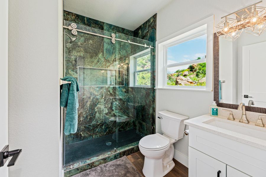 Bathroom featuring a striking green stone shower enclosure with a glass door, a toilet, a window, a single vanity with a white countertop and undermount sink, and wood-finish flooring