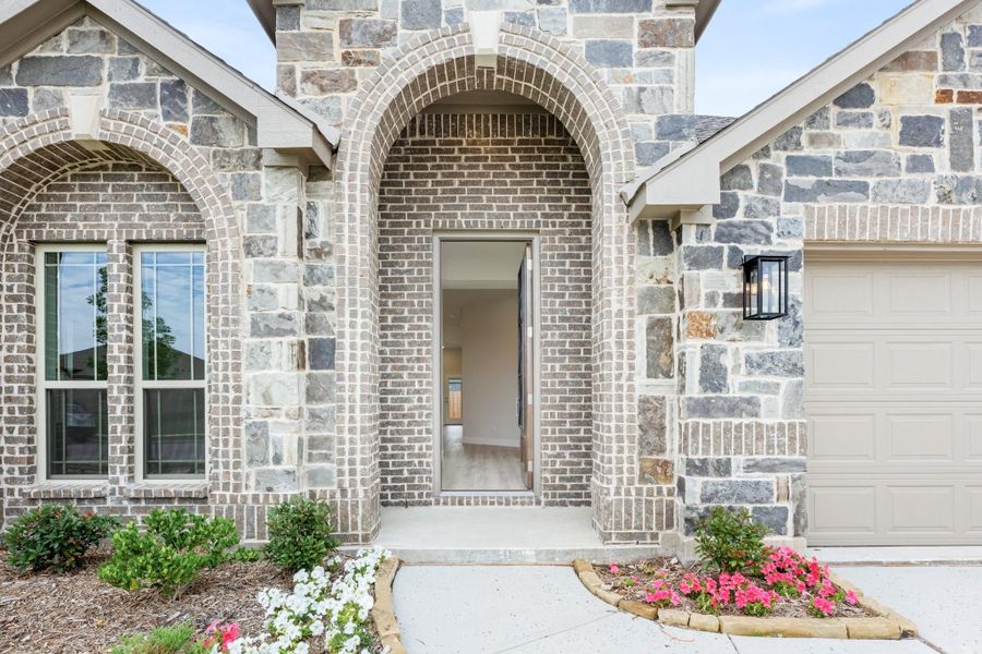 Home entrance with brick and stone exterior, arched doorway, wall sconce, and landscaped flower beds near garage