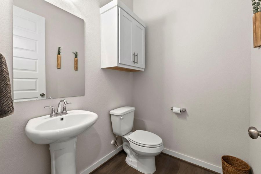 Pedestal sink with chrome faucet, a rectangular wall-mounted mirror, white paneled door, wood-finish flooring, and a wall-mounted white cabinet with chrome hardware