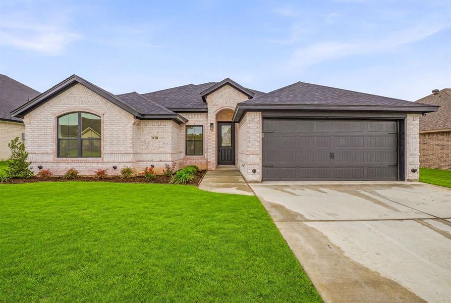 The property features a light-colored brick exterior, a dark shingle roof, a well-maintained green lawn, and a concrete driveway leading to a dark-colored garage door