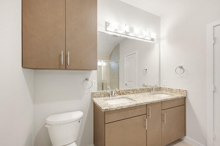 Bathroom with double sink vanity, granite countertop, brown cabinets, large mirror, and toilet