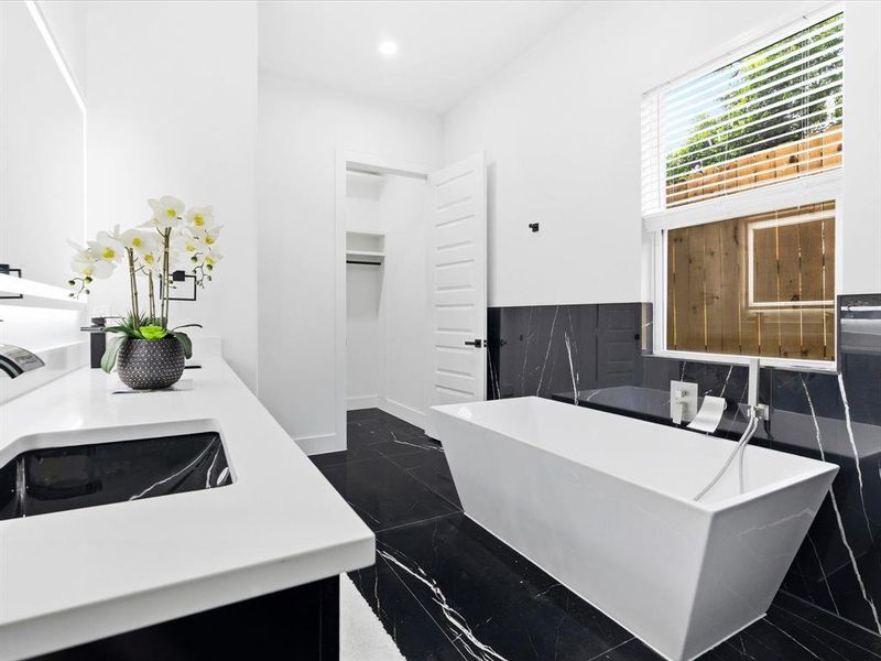 Modern bathroom featuring a freestanding soaking tub, a single vanity with an integrated sink, black and white veined tile flooring, a horizontal paneled window with blinds, and a built-in storage closet