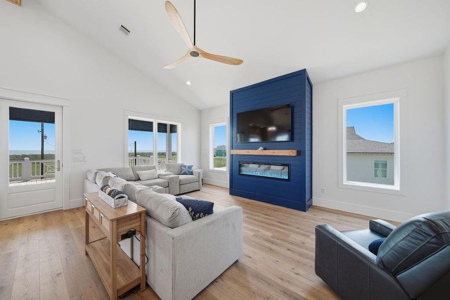 Bright and spacious living room with high ceilings, large windows, and wood flooring. Features a modern blue accent wall with a mounted TV and an electric fireplace. Cozy seating area and access to outdoor views.