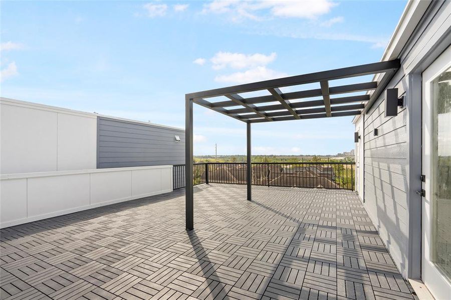 This is a spacious, modern rooftop patio featuring herringbone-patterned floor tiles, a sleek metal pergola, and a high privacy fence, offering an open-air entertainment space with a clear view of the surrounding landscape.