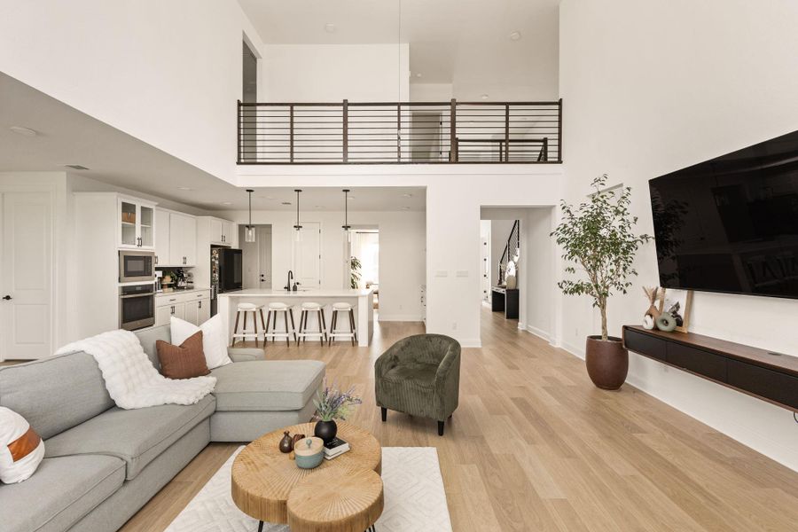 Open-concept living area with soaring ceilings, light wood-finish flooring, and a modern kitchen featuring white cabinetry, a large island with seating, and stainless steel appliances