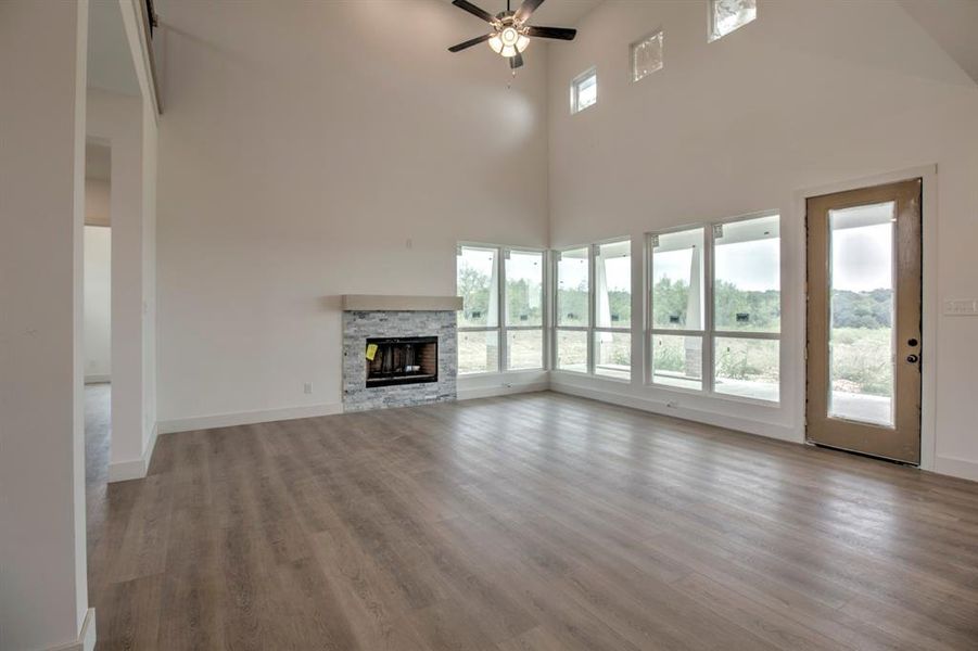 Unfurnished living room with a stone fireplace, light wood-type flooring, a towering ceiling, and a ceiling fan Unfurnished living room with a stone fireplace, light wood-type flooring, a towering ceiling, and a ceiling fan