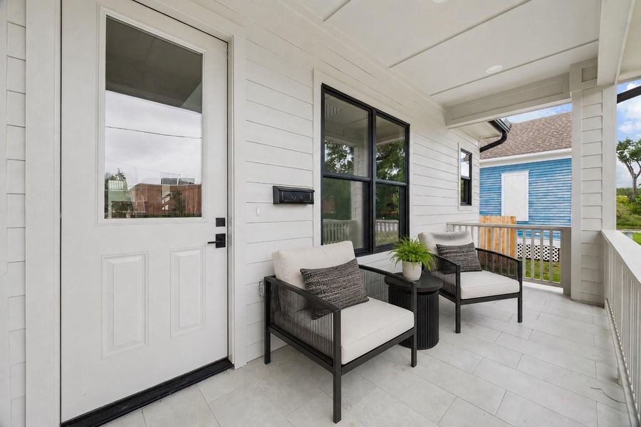 The covered front porch creates an inviting outdoor sitting area overlooking the surrounding neighborhood and park-adjacent greenery. Contemporary lighting, black-framed windows, and modern staging elements complement the home’s clean exterior palette and indoor-outdoor feel. (digitally staged)
