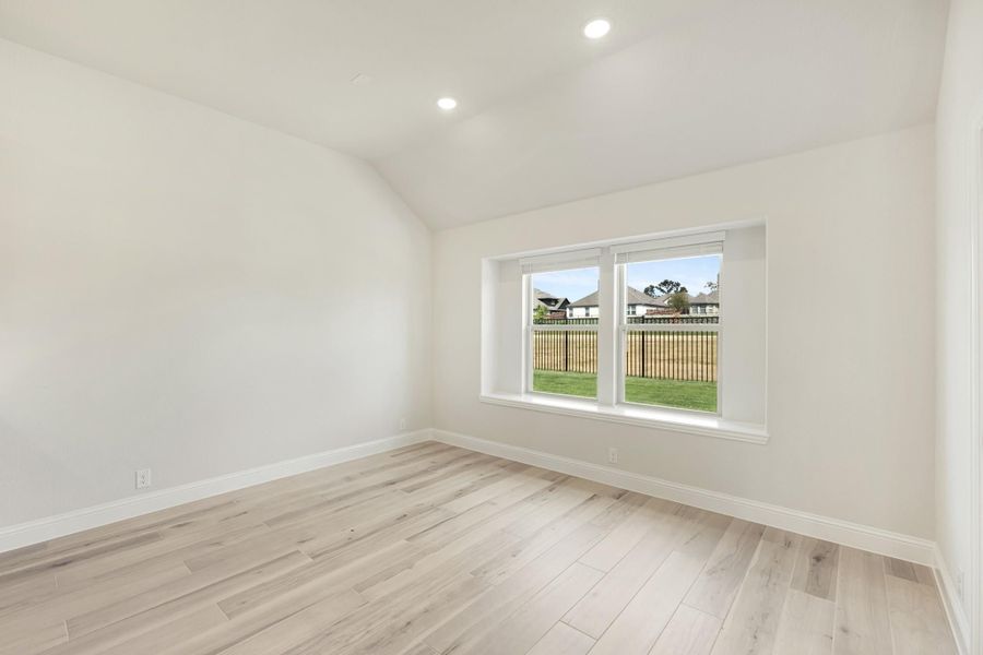 Empty bedroom with vaulted ceiling, light wood floors, and a window overlooking a fenced backyard