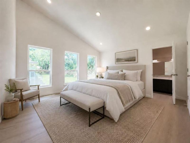 Spacious bedroom featuring a vaulted ceiling with recessed lighting, three large windows, light wood-finish flooring, and direct access to a bathroom with a dark wood-finish vanity Spacious bedroom featuring a vaulted ceiling with recessed lighting, three large windows, light wood-finish flooring, and direct access to a bathroom with a dark wood-finish vanity