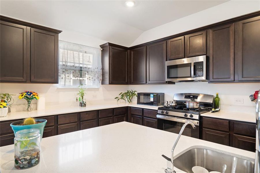 Kitchen with dark brown cabinets, appliances with stainless steel finishes, lofted ceiling, and light stone countertops
