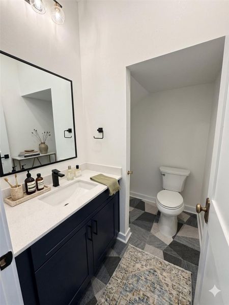 This is a stylish, modern bathroom with a navy vanity, white countertop, and geometric floor tiles. It features a sleek mirror, contemporary lighting, and a separate toilet area.