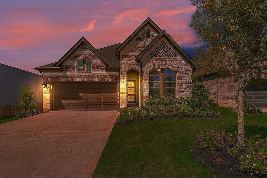 Charming brick and stone exterior home with a well-maintained front yard, illuminated by warm lighting. Features a double garage and a welcoming entrance, set against a picturesque sunset sky.