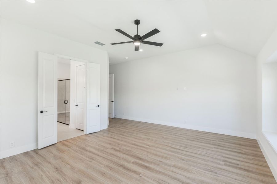Empty bedroom with light wood floors, vaulted ceiling, black ceiling fan, and open doors to ensuite bathroom
