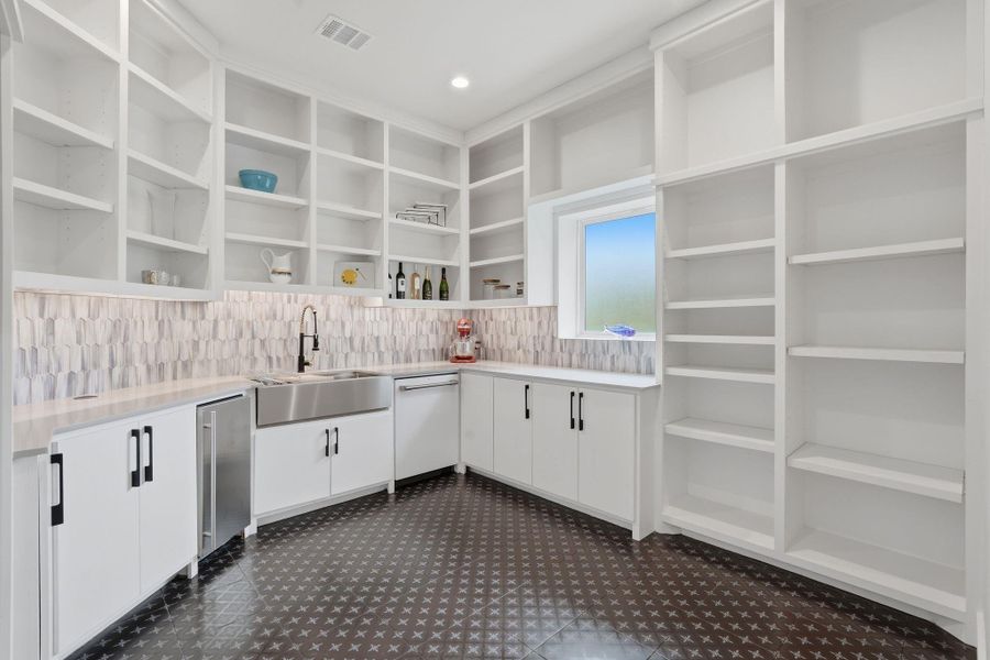 This is a bright, spacious walk-in butlers pantry with ample open shelving for storage, a modern farmhouse sink, stylish backsplash, and sleek cabinetry. The room features a patterned tile floor and a window for natural light, hidden behind the pantry doors in the main kitchen.
