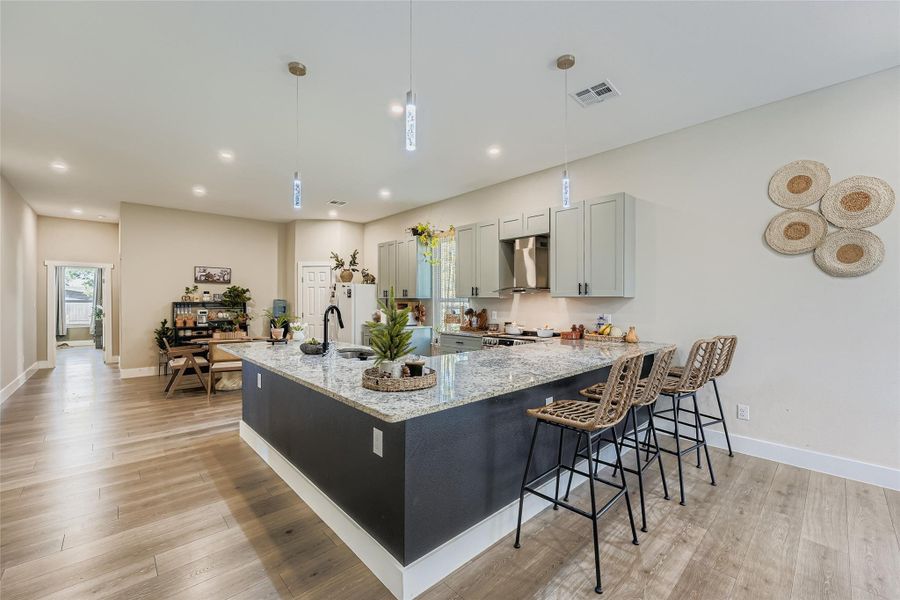 Kitchen featuring light stone countertops, a kitchen breakfast bar, decorative light fixtures, gray cabinets, and wall chimney exhaust hood Kitchen featuring light stone countertops, a kitchen breakfast bar, decorative light fixtures, gray cabinets, and wall chimney exhaust hood