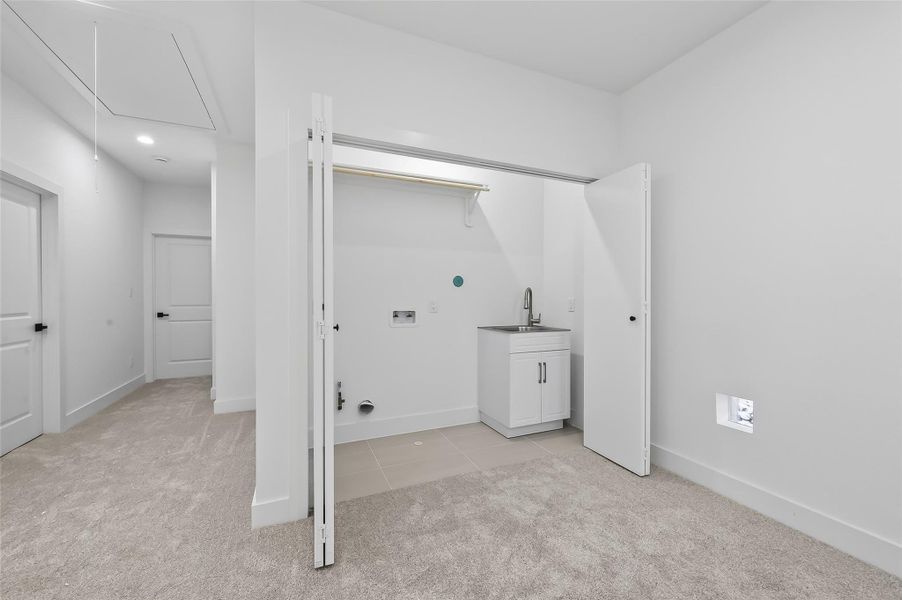 This photo shows a bright, laundry room with fresh white walls and carpeted flooring. It features a utility sink, washer/dryer hookups, and a storage shelf. The space is well-lit and adjacent to a hallway, providing easy access.