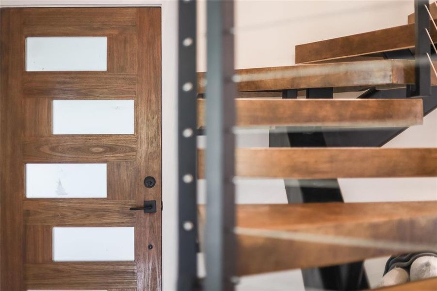 A modern wooden entrance door features frosted glass panels next to a striking open staircase.