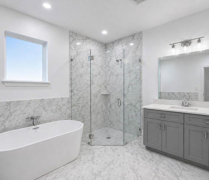 Spacious bathroom featuring a freestanding soaking tub, a frameless glass shower enclosure, a dual-sink vanity with gray cabinetry and white countertops, and marble-patterned tile flooring and shower surround