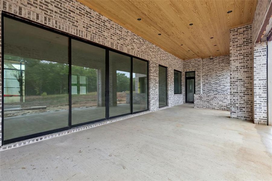 Spacious covered patio featuring a concrete floor, brick walls, a wood-finish ceiling with recessed lighting, and expansive multi-panel sliding glass doors
