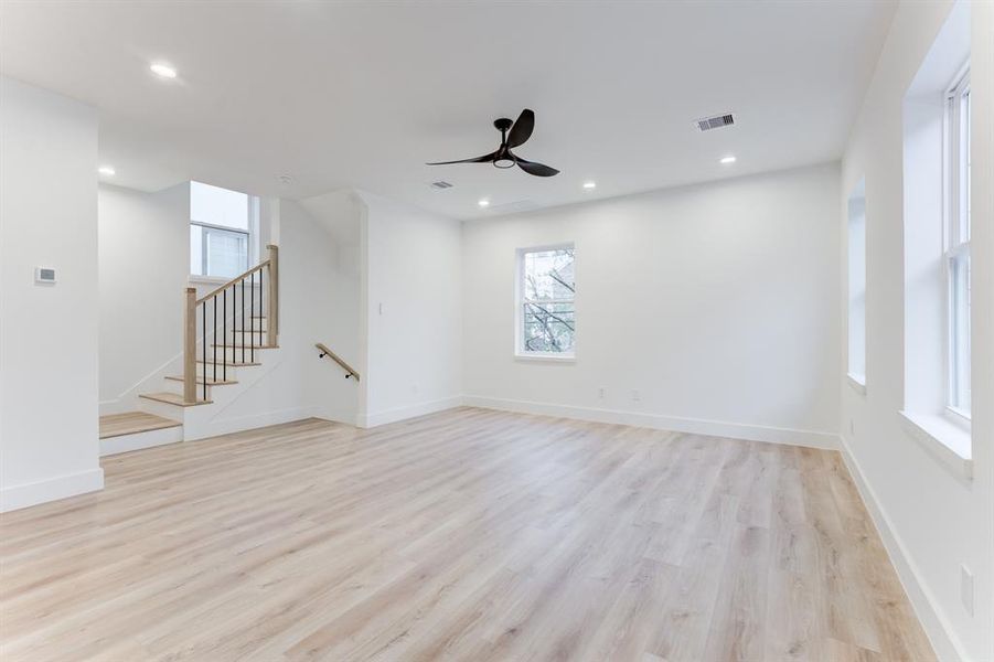 This spacious living room features an open layout with light wood flooring, modern decor, and ample natural light from large windows, ceiling fan, and recessed lighting.