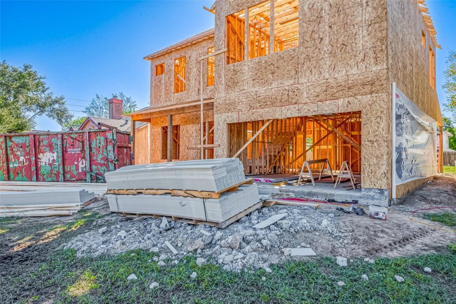 This image shows a house under construction, featuring exposed wooden framing and exterior sheathing. Construction materials are stacked in the yard, and a large dumpster is nearby.