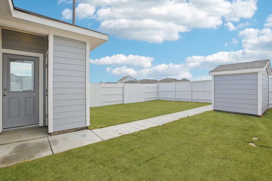Image of a light grey house back exterior with a small grey shed, green grass, a white fence, and a blue sky in the background Image of a light grey house back exterior with a small grey shed, green grass, a white fence, and a blue sky in the background