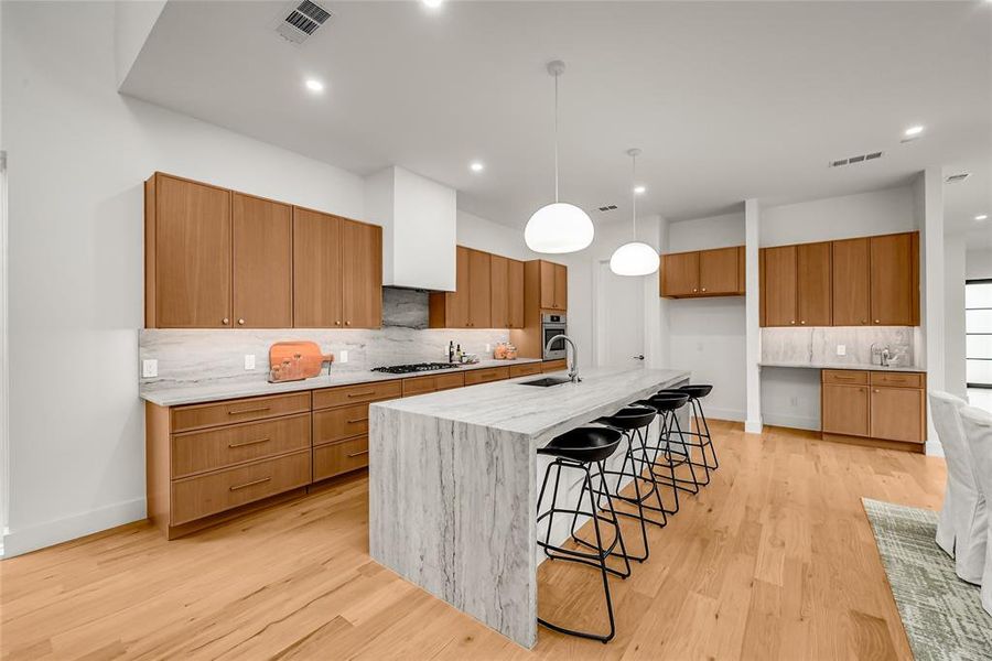 Contemporary kitchen featuring a waterfall island with integrated seating, light wood cabinetry, stone countertops, and wood-finish flooring