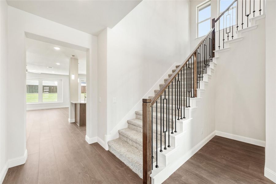 Carpeted staircase with wood handrail and iron balusters in an open entryway with hardwood floors