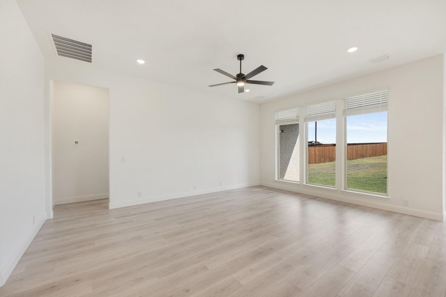 Empty living room with light wood floors, ceiling fan, and large windows overlooking backyard