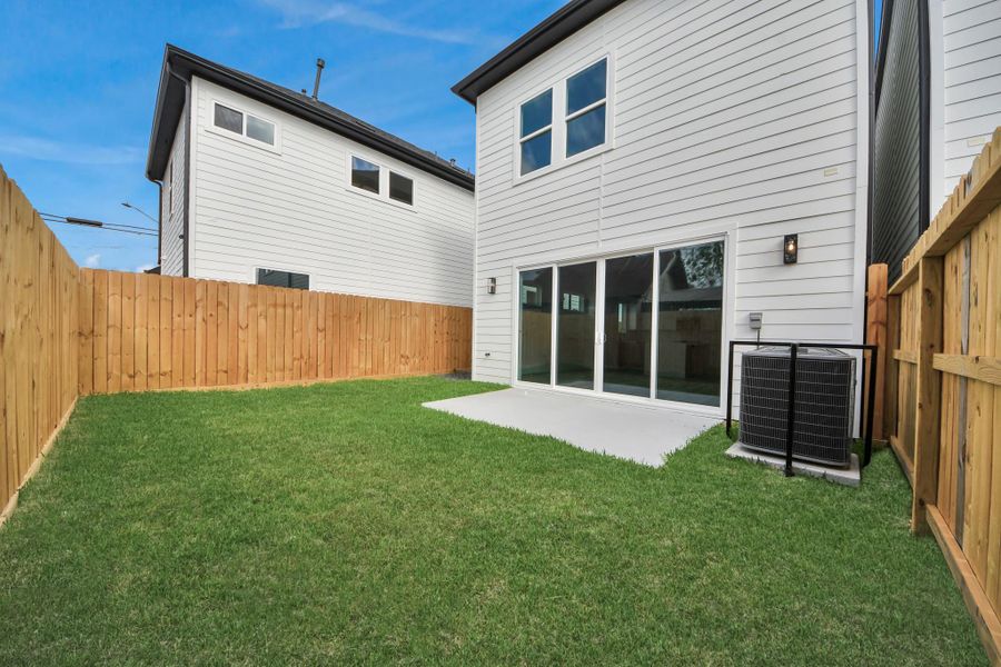 This photo shows a modern two-story house with a well-maintained, fenced backyard featuring a small lawn and a concrete patio area. The exterior is clad in white siding, and there are large sliding glass doors leading into the home.