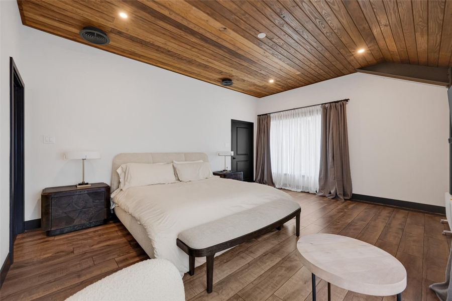 Wood-paneled ceiling, hardwood floors, and a large window with sheer curtains. It includes a comfortable bed, modern side tables with lamps, and a small seating area, creating a warm and inviting space.