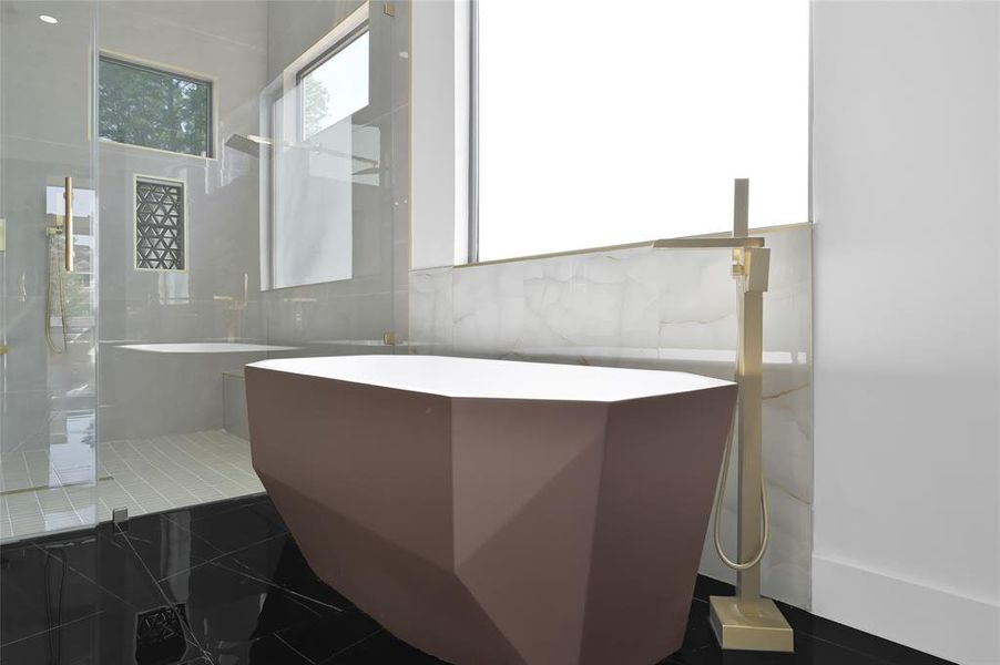 This bathroom features a sleek, modern design with a geometric freestanding bathtub, many windows for natural light, and a luxurious glass-enclosed shower with gold fixtures that add an elegant touch.