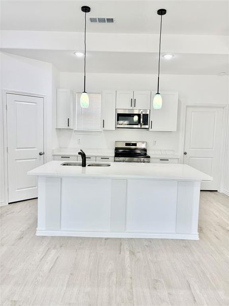 Kitchen featuring white cabinetry, hanging light fixtures, an island with sink, and light stone countertops