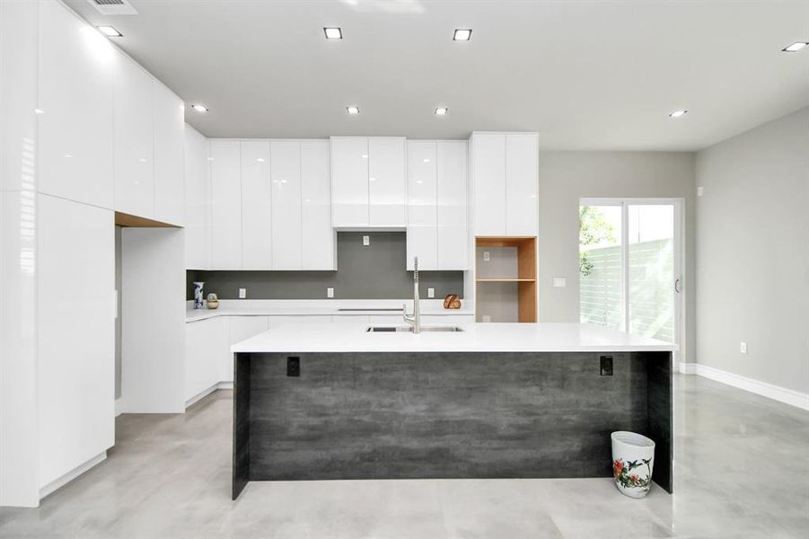 Modern kitchen with sleek white cabinetry, a large island with a dark finish, built-in shelves, and abundant natural light from a glass door leading to an outdoor space.