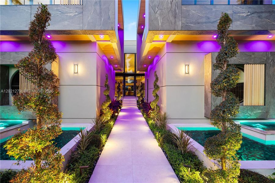 Resort-style entry corridor with dual reflecting pools, spiral topiaries, wood-paneled ceiling, recessed lighting, and illuminated pathway leading to glass foyer entrance.