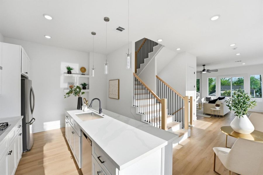 Open-concept kitchen and living area featuring a spacious quartz island with seating, white shaker cabinetry, pendant lighting, wood-look flooring, and a striking modern staircase that enhances the home’s bright, contemporary design.