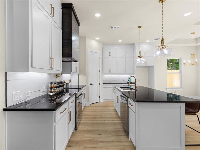 Gourmet kitchen featuring white shaker cabinetry with brass hardware, dark countertops, wood-finish flooring, and a large center island with built-in sink