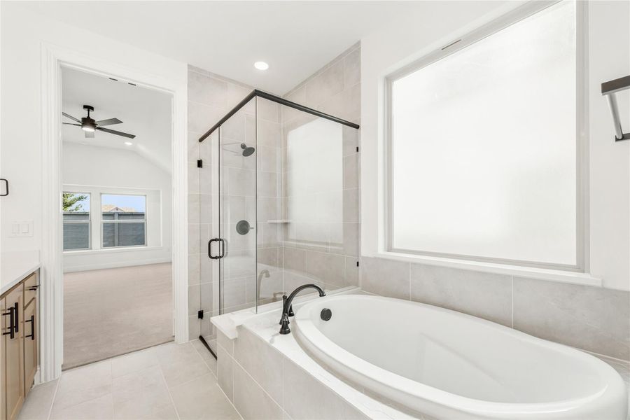 Master bathroom with soaking tub, glass-enclosed shower, gray tile, and view into bedroom with ceiling fan