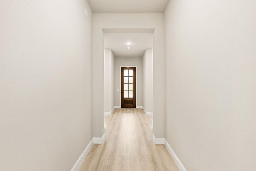 Narrow hallway with light wood floors, white walls, and dark wood glass-panel front door at the end Narrow hallway with light wood floors, white walls, and dark wood glass-panel front door at the end
