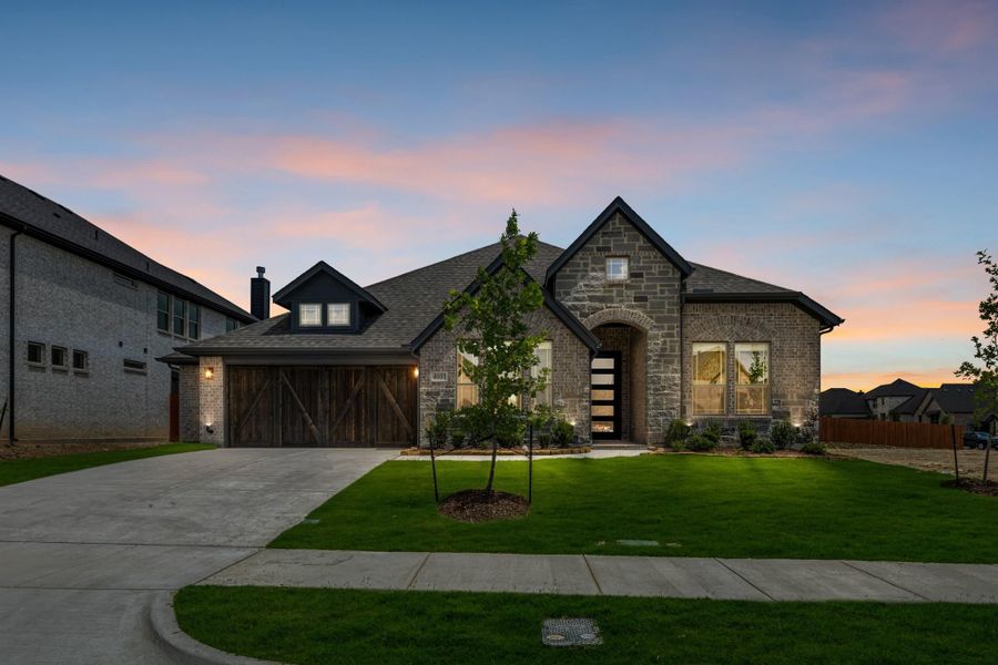 Single-story stone and brick home with wood garage door, arched entry, and green front lawn at dusk. Single-story stone and brick home with wood garage door, arched entry, and green front lawn at dusk.