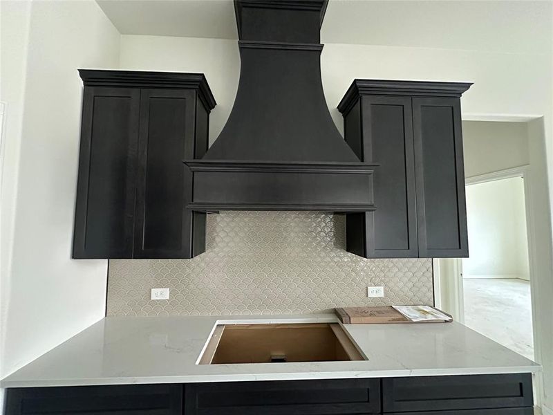Kitchen with custom exhaust hood, dark cabinets, light stone countertops, and tasteful backsplash