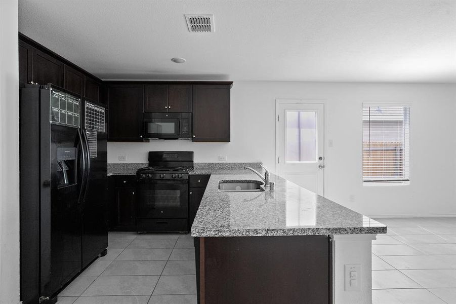 Kitchen with black appliances, light tile patterned floors, a peninsula, light stone counters, and dark brown cabinets
