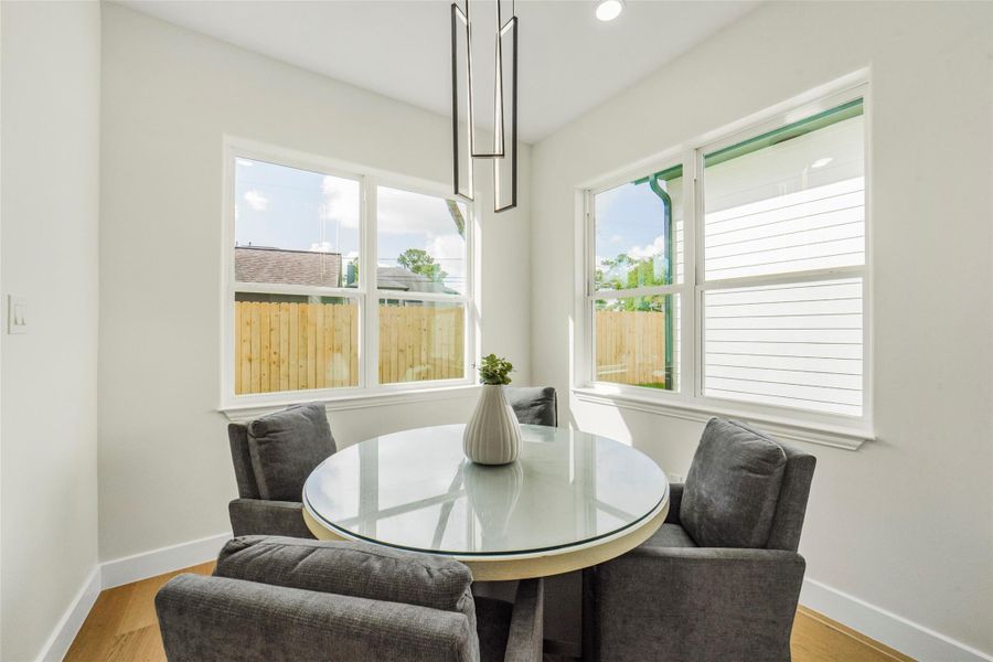 This bright dining area features a round glass table with four cushioned chairs, surrounded by large windows that offer plenty of natural light and views of a fenced backyard.