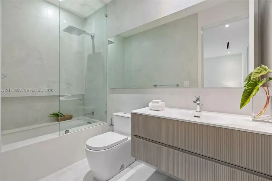 Floating vanity with ribbed cabinetry and integrated sink, frameless glass shower enclosure, rainfall showerhead, large wall-spanning mirror, and neutral-toned wall tiling