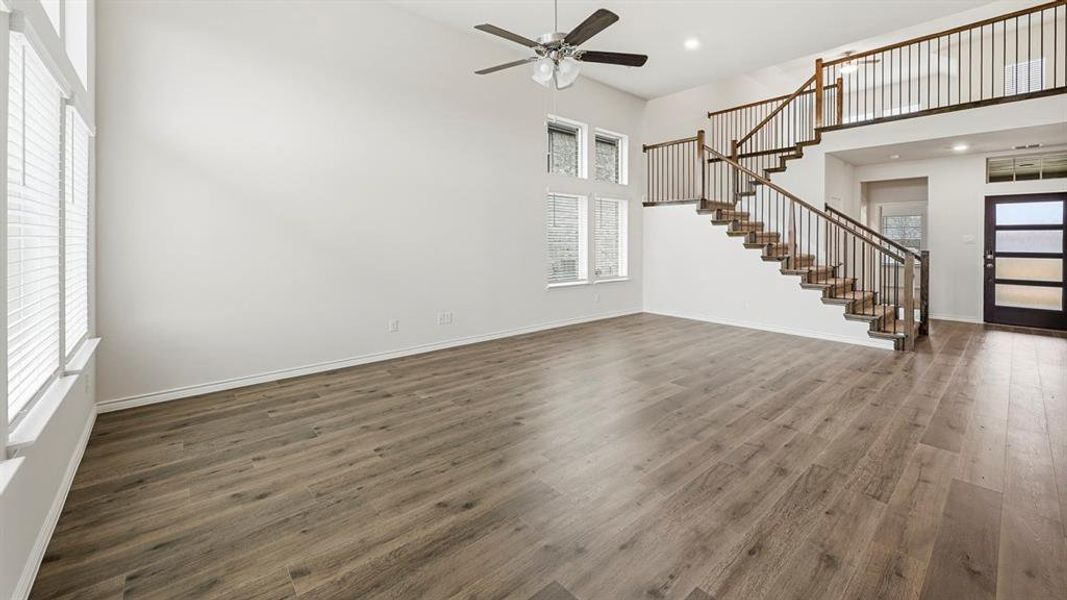 Spacious living area featuring high ceilings, wood-finish flooring, a contemporary ceiling fan, multiple large windows, and a prominent staircase with dark wood handrails and balusters
