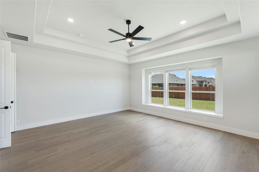 Empty bedroom with tray ceiling, ceiling fan, hardwood floors, and large window overlooking backyard Empty bedroom with tray ceiling, ceiling fan, hardwood floors, and large window overlooking backyard
