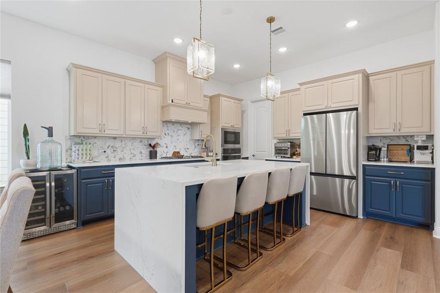 Modern kitchen featuring a large central island with seating, two-tone cabinetry, stainless steel appliances, wood-finish flooring, and pendant lighting Modern kitchen featuring a large central island with seating, two-tone cabinetry, stainless steel appliances, wood-finish flooring, and pendant lighting
