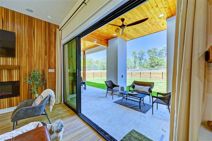 This inviting home features a seamless indoor-outdoor living space with large sliding glass doors leading to a covered patio. The interior boasts a modern, wooden accent wall and cozy seating, while the patio offers relaxed outdoor furniture and views of a fenced, tree-lined yard. Perfect for entertaining and enjoying nature.