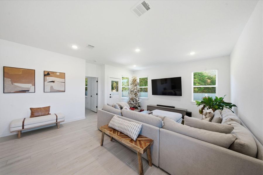 This spacious living room features a large sectional sofa, a mounted TV, and abundant natural light from multiple windows. The neutral tones and minimalist decor create a welcoming and modern atmosphere.