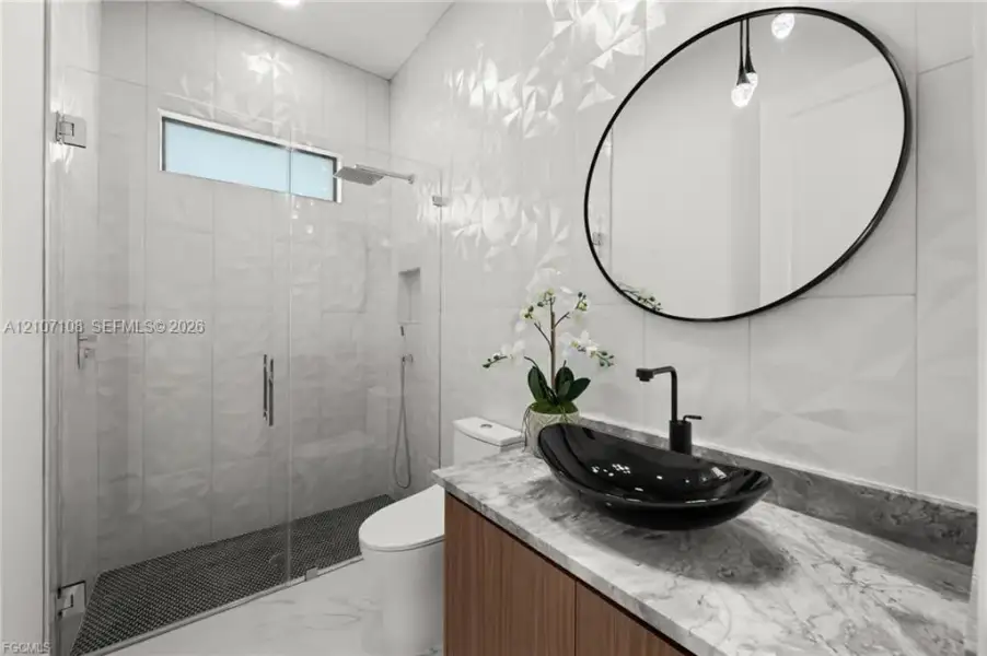 Bathroom featuring a custom wood-finish vanity with a stone-top surface, black vessel sink, geometric textured wall tile, frameless glass shower enclosure, and a circular black-framed mirror